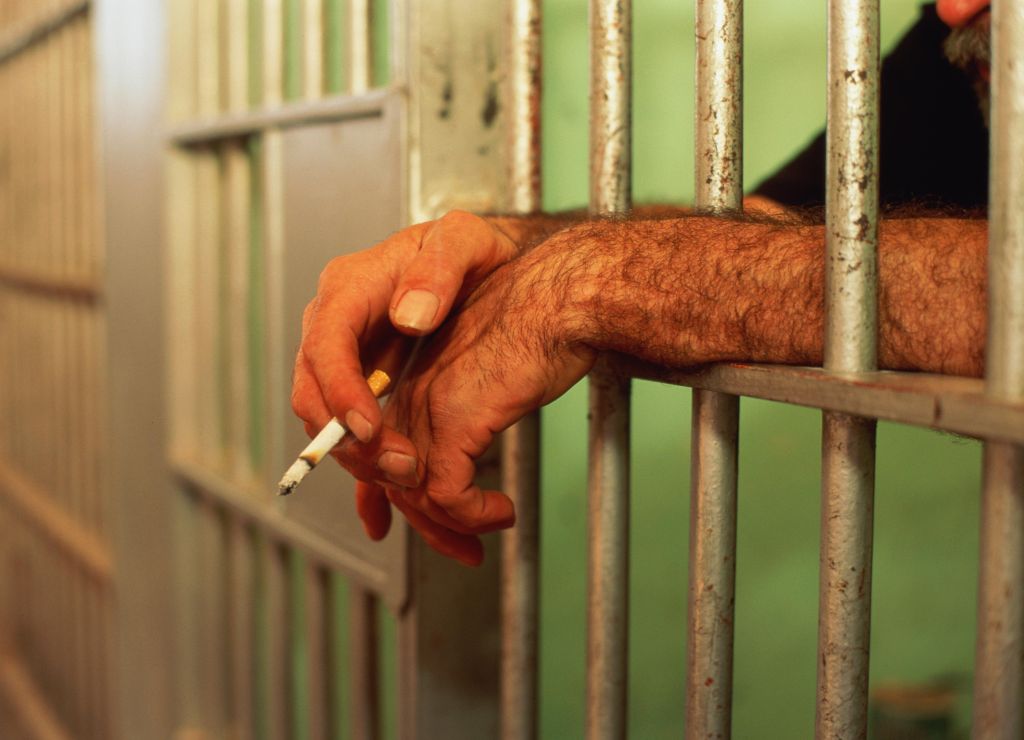 Man's hands poking through bars of jail cell, holding lit cigarette
