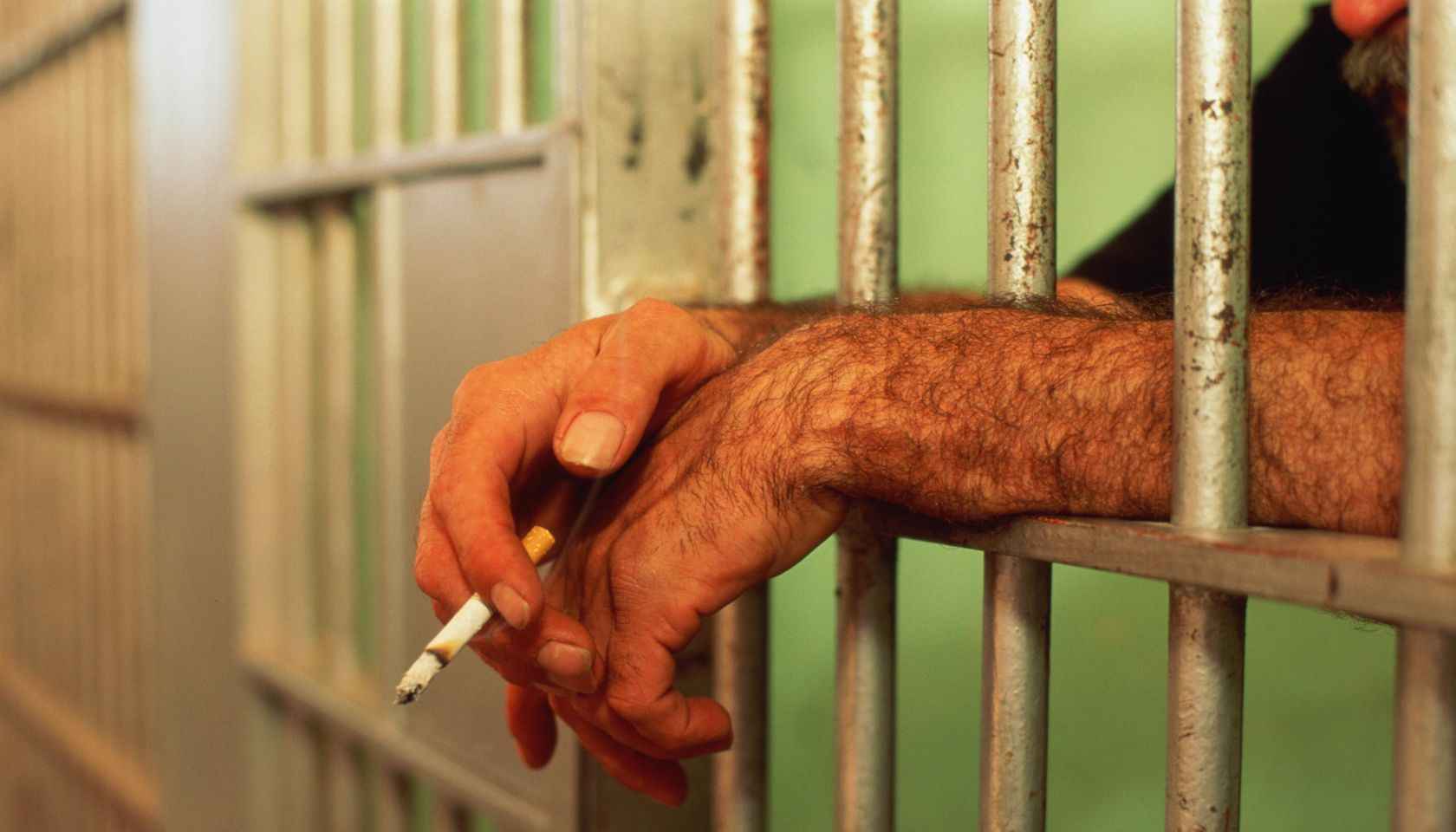 Man's hands poking through bars of jail cell, holding lit cigarette