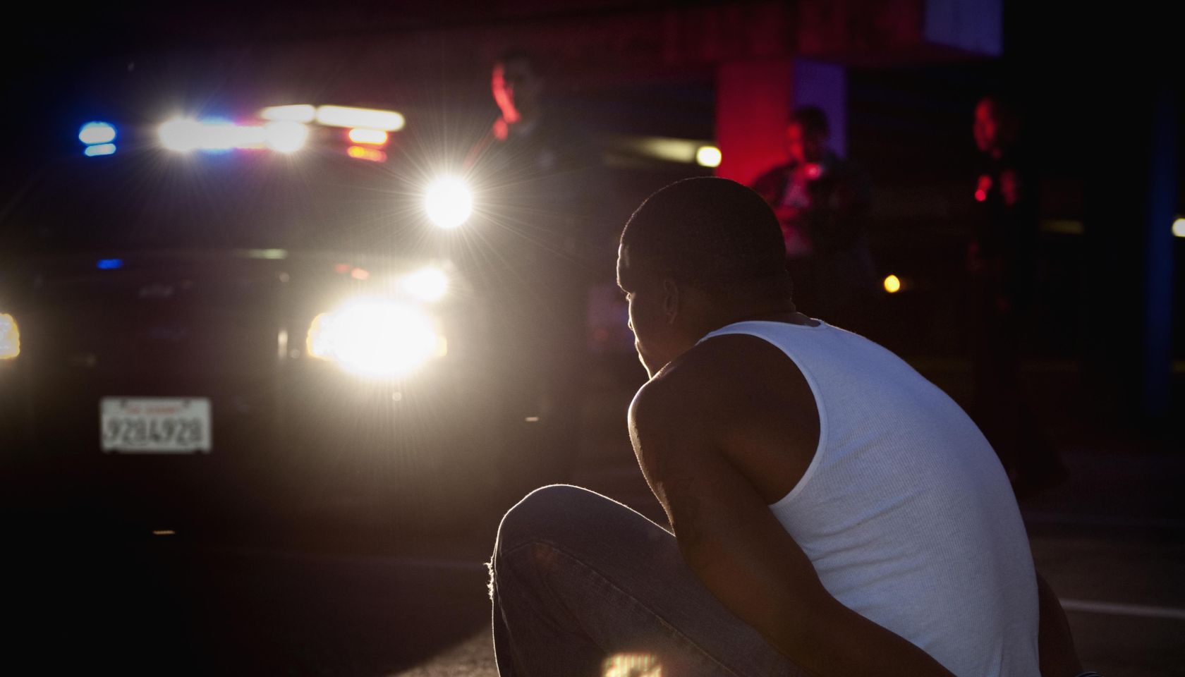 Police shining lights on handcuffed African man sitting on curb