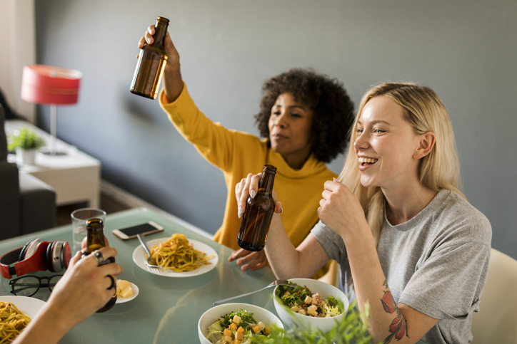 Happy girlfriends sitting at dining table raising beer bottles