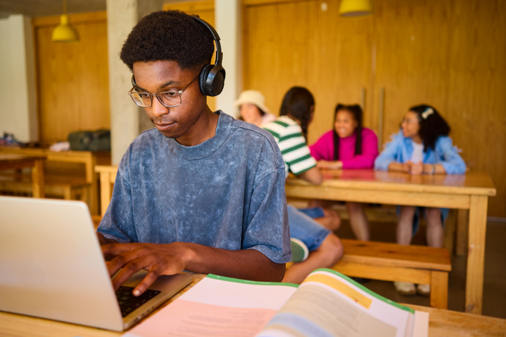 Focused teen studying on laptop with headphones in library, while friends socialize