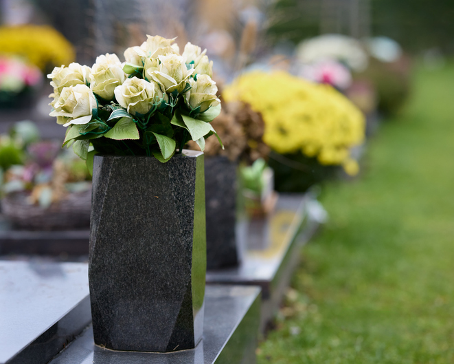 Artificial white roses in a dark granite vase on a tombstone, with blurred graves and green grass in the background, creating a solemn memorial scene.