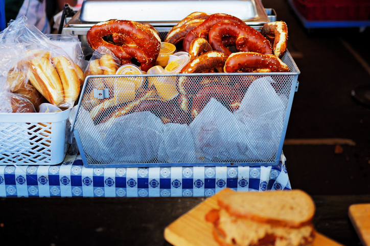 Assortment of freshly baked soft pretzels and bread rolls displayed at an outdoor market stall, with a sandwich in the foreground