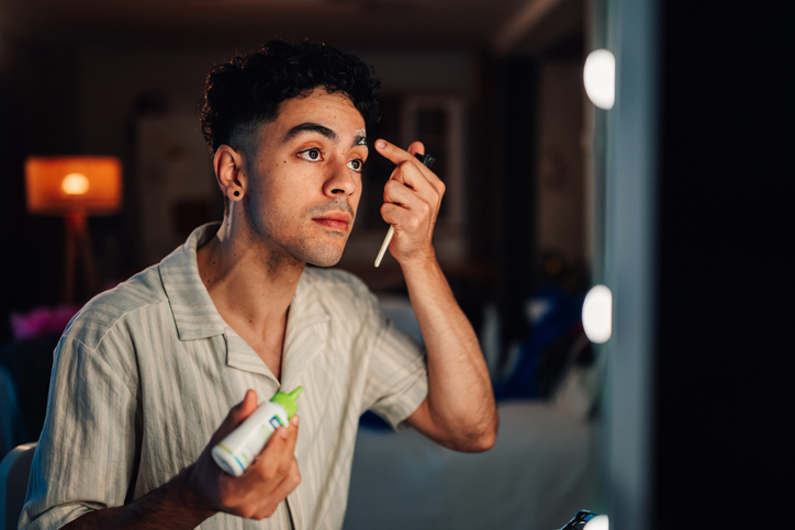 Young man applying makeup to his eyebrow in a dressing room mirror, preparing for a performance or self-care routine