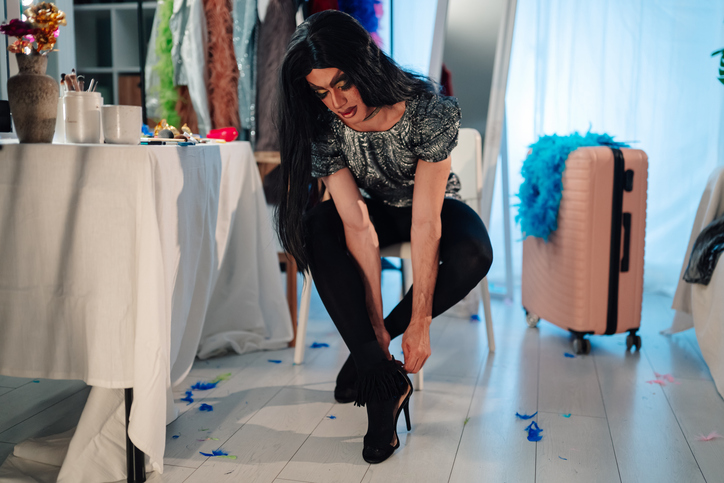 Drag performer putting on high heels, preparing for a show in a vibrant backstage dressing room