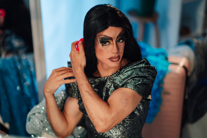 Drag queen preparing for performance, applying wig in dressing room showcasing lgbtq+ art and culture