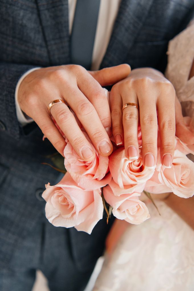 Close-up of bride and groom hands with golden wedding rings and flower bouquet.