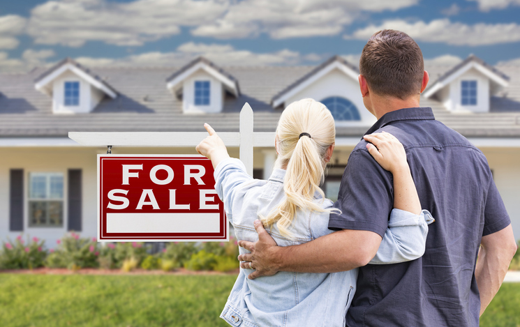 Young adult couple facing and pointing to front of for sale real estate sign and house