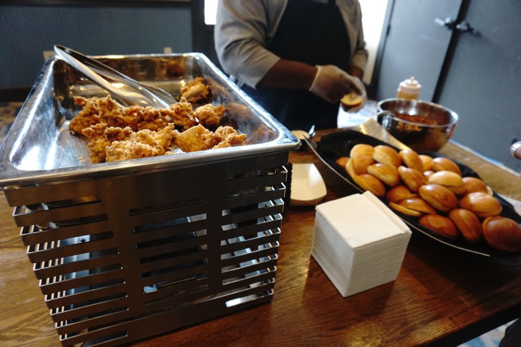 A tray of fried chicken and rolls on a wooden table, with a person's hand visible in the background.