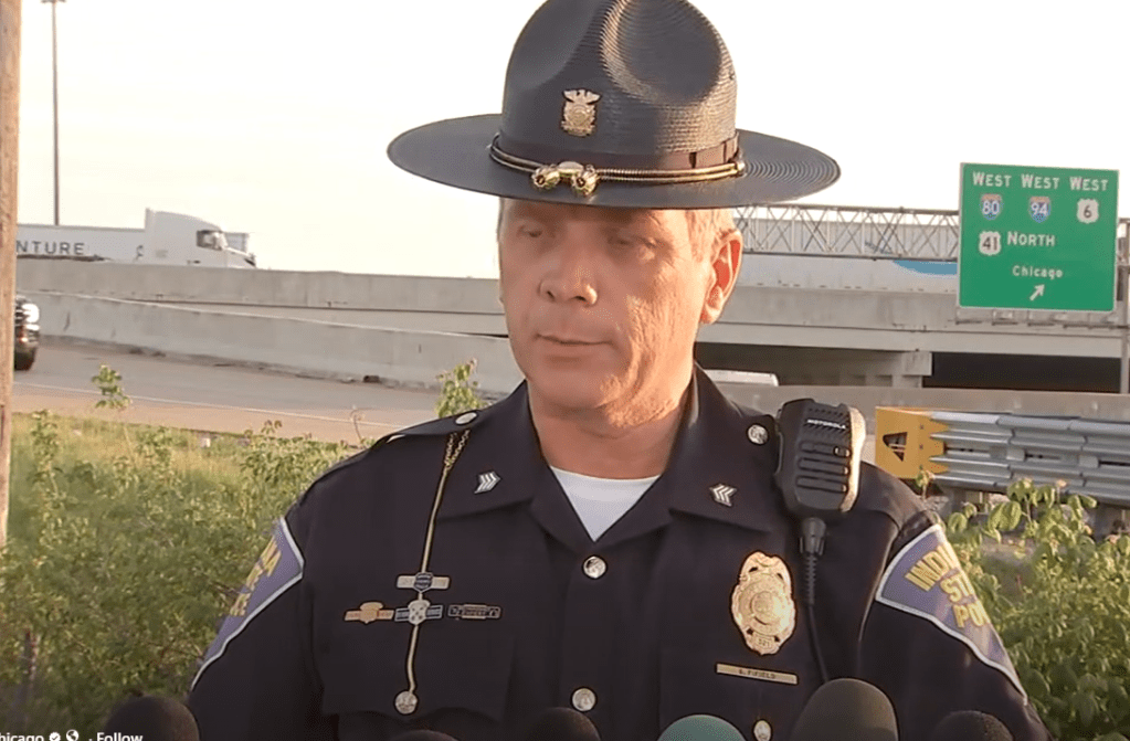 A uniformed police officer standing in front of a highway sign, speaking into a radio.