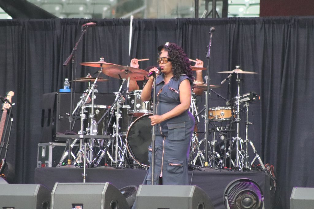 A woman with curly hair singing into a microphone on a stage with drums and other musical equipment behind her.