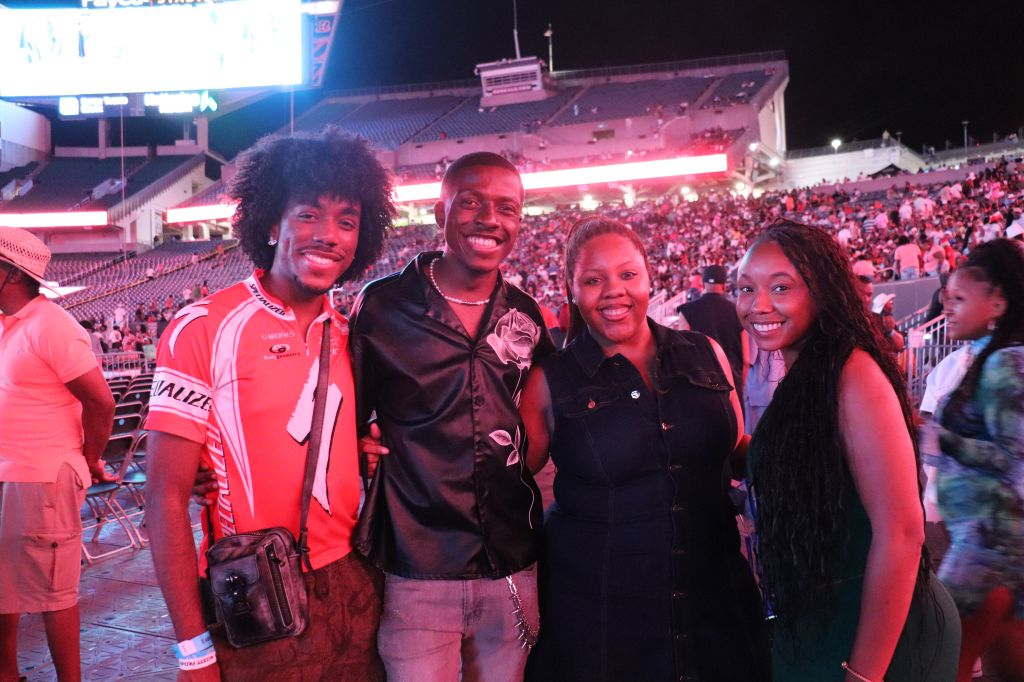 A group of four smiling people, including two men and two women, posing together in a crowded stadium at night.