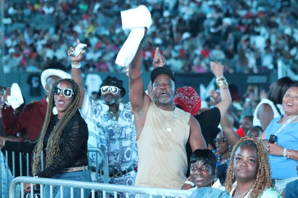 Crowd of people at an event, some raising their hands and holding signs, with a person in the center wearing a white shirt and hat.