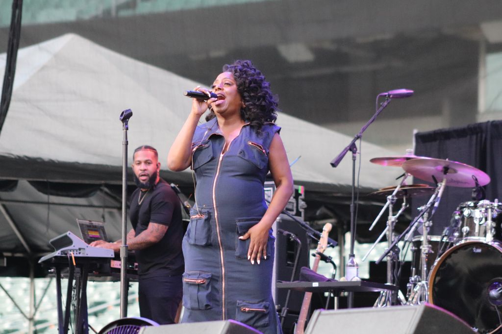 A woman with curly purple hair singing into a microphone on stage, accompanied by a man playing a keyboard.