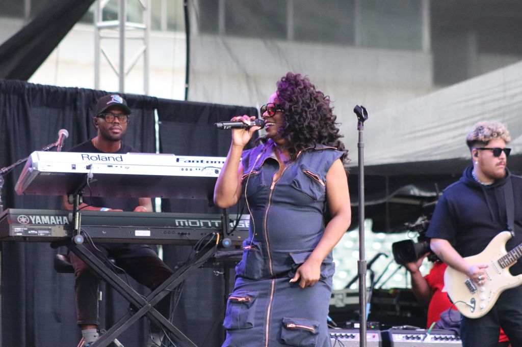 A musician with curly purple hair performing on stage, playing a Roland keyboard and singing into a microphone, with a guitarist and drummer in the background.