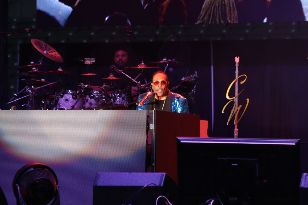 A musician performing on stage, wearing sunglasses and a colorful sequined jacket, playing a keyboard with a large logo behind them.