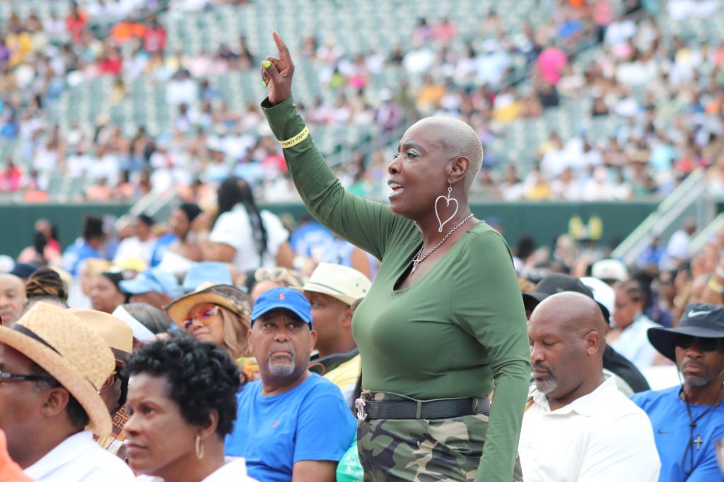 A woman with a shaved head raises her hand while addressing a large crowd of people at an outdoor event.