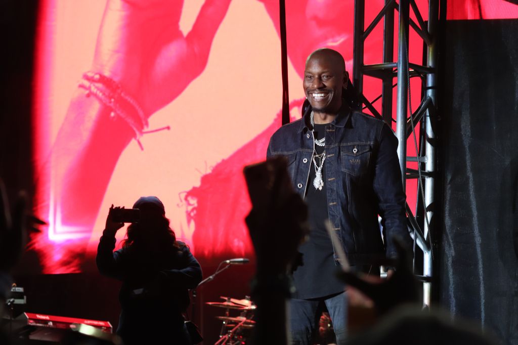A smiling man wearing a denim jacket stands on a stage with a large red backdrop.