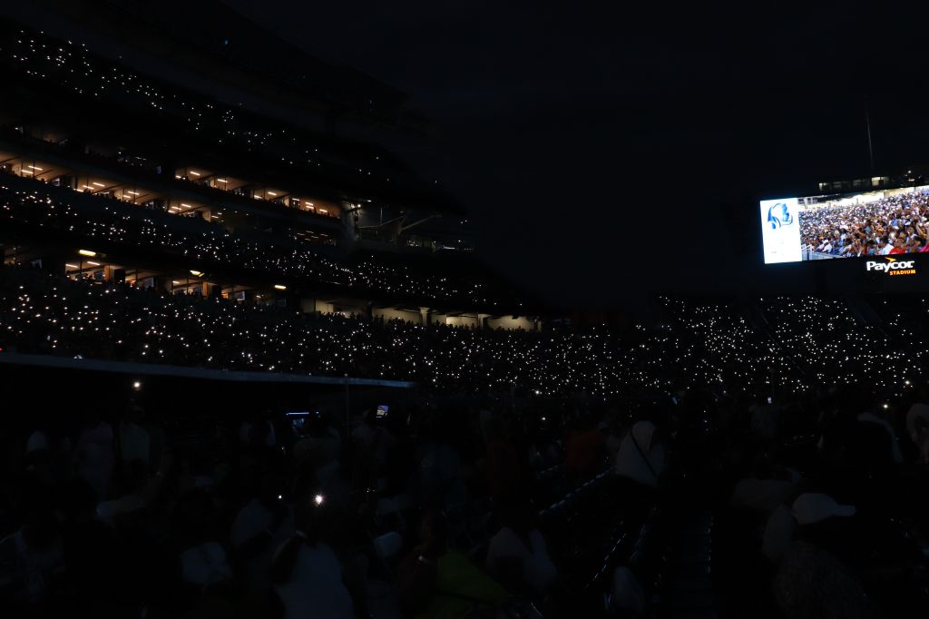 A large crowd of people holding up their phones in a darkened stadium, creating a sea of lights against the night sky.