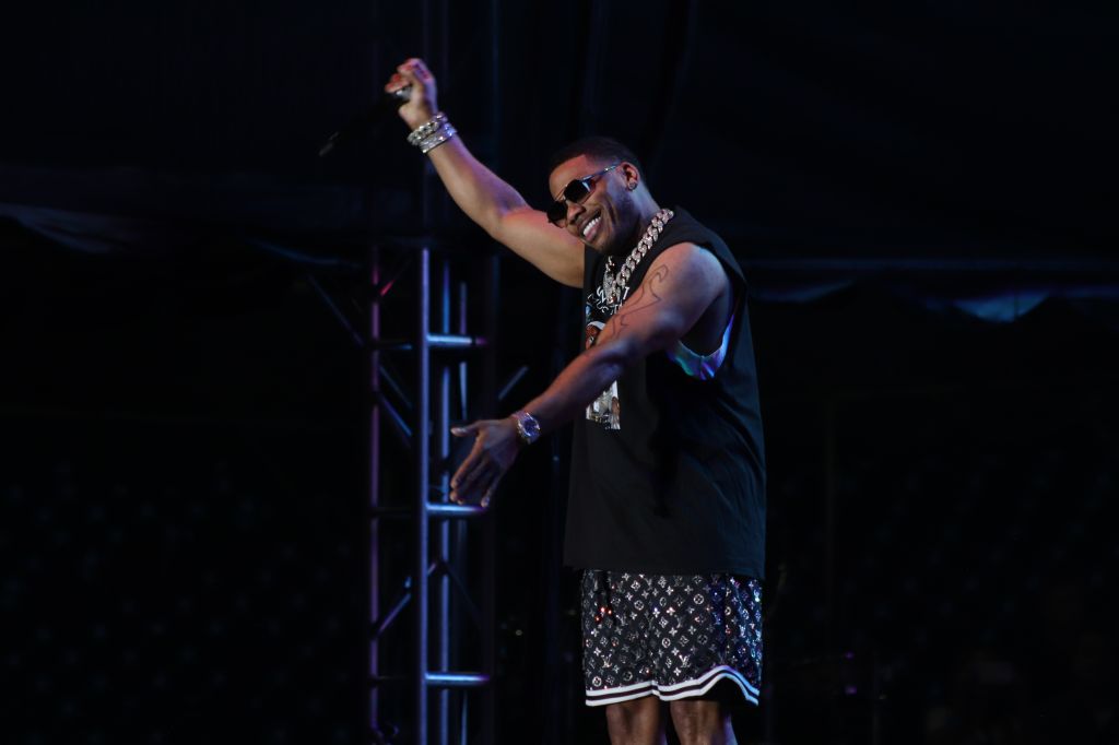 A performer on stage wearing sunglasses, jewelry, and a patterned outfit, raising one arm in a celebratory gesture.