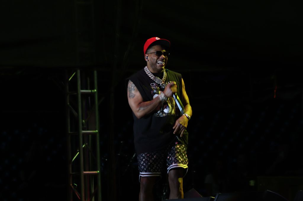A smiling man wearing a red cap, black shirt, and yellow jacket performing on a stage in a dark setting.