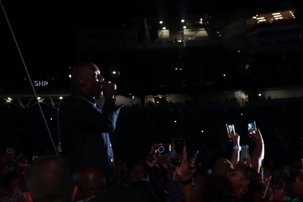 A person speaking on stage in a dark venue, with the audience visible in the background holding up their phones.