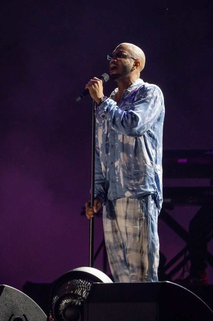A man wearing a tie-dye shirt and glasses singing into a microphone on stage in a dimly lit venue.