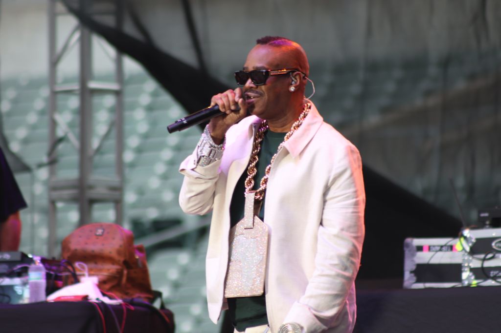 A man wearing sunglasses and jewelry performs on stage, holding a microphone and wearing a white jacket.