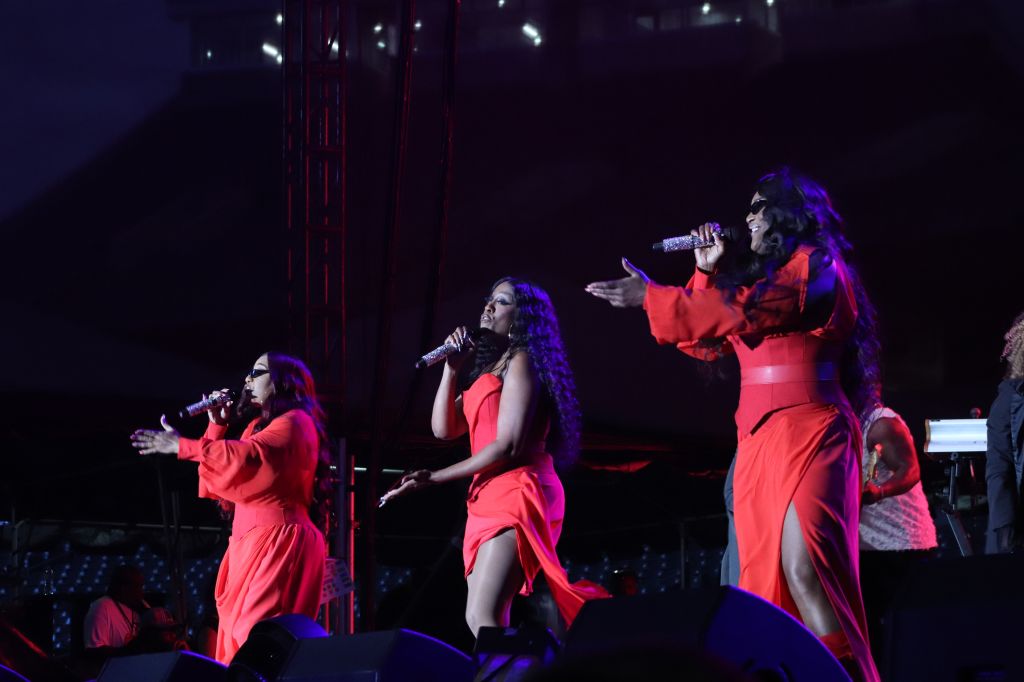 Three female singers performing on stage in red dresses, singing into microphones.