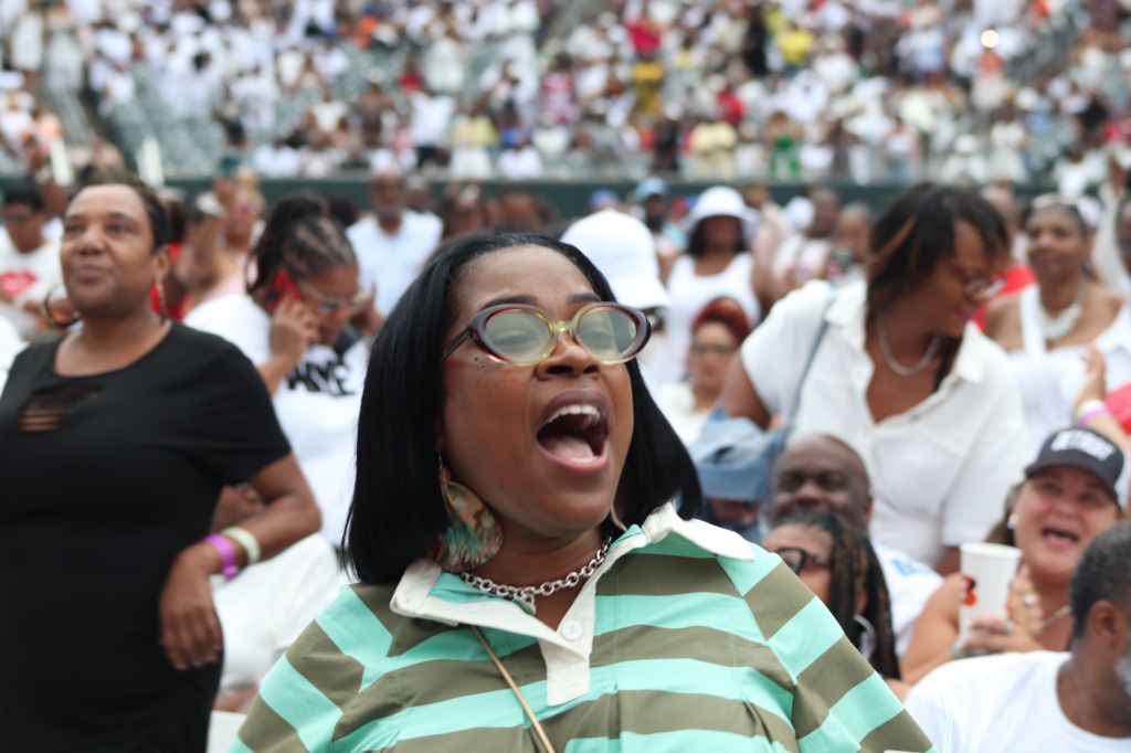 A large crowd of people, many wearing colorful clothing, with a woman in the foreground shouting with her mouth open wide and wearing round glasses.