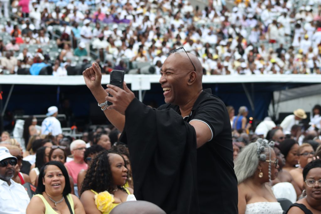 A smiling man in a black shirt raises his fist while surrounded by a large crowd at an event.
