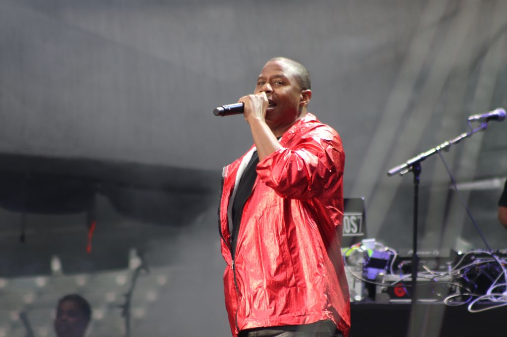 A man in a red jacket singing into a microphone on stage with musical equipment visible behind him.