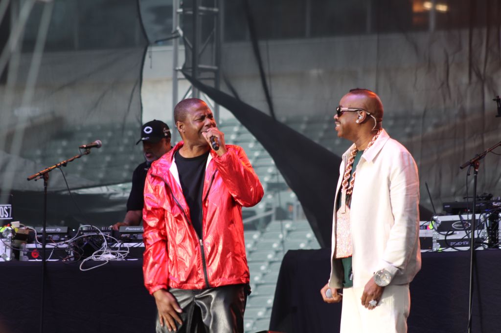 Two men performing on stage, one wearing a red jacket and the other a white suit, with musical equipment and a backdrop visible.