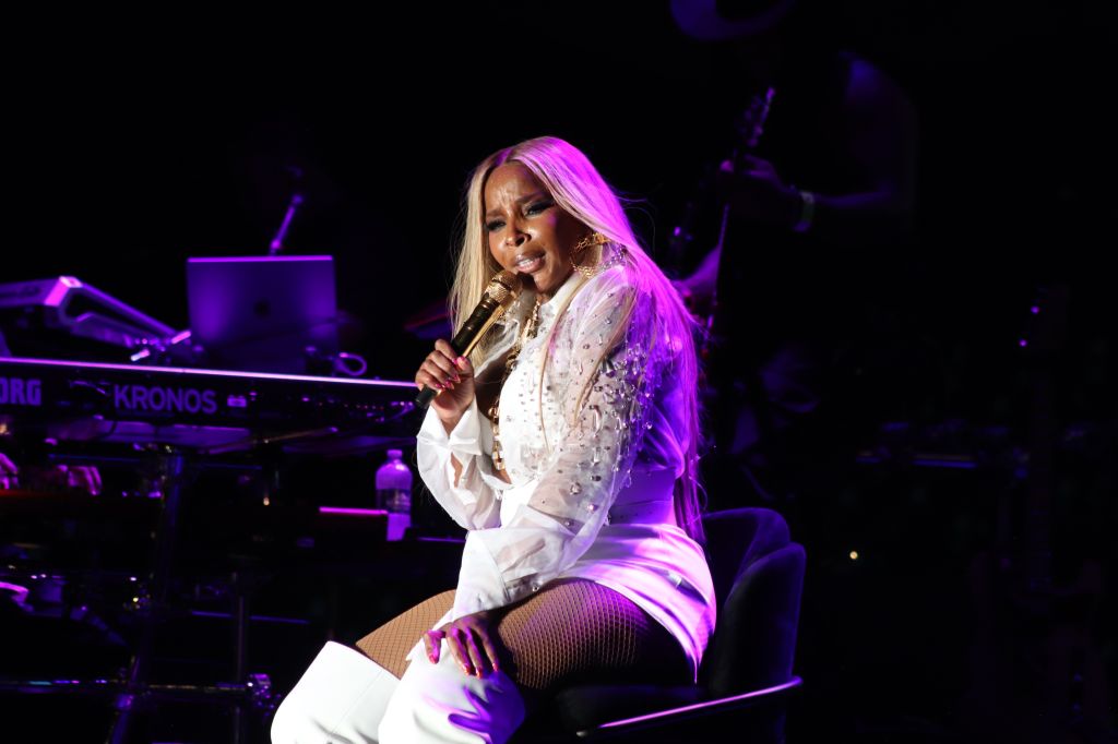 A woman with long blonde hair performing on stage, holding a microphone and wearing a white outfit with sparkling embellishments. The stage is illuminated with purple lighting.