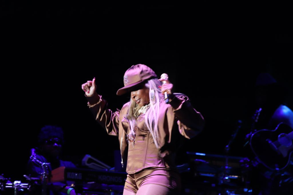 A performer wearing a pink outfit and hat dancing on stage in a dimly lit concert setting.